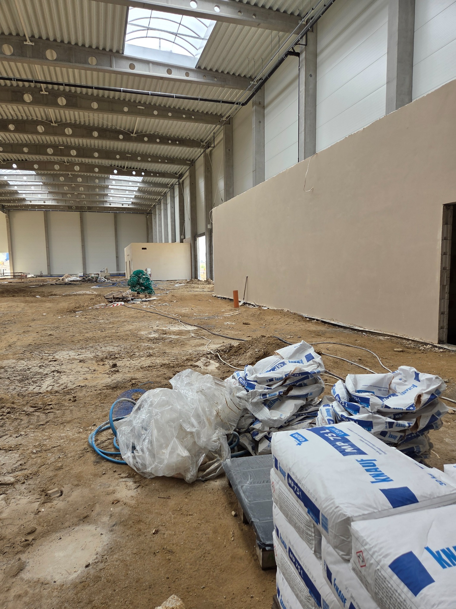 Wnętrze hali w trakcie prac tynkarskich: fragment ściany z nałożonym tynkiem, worki z zaprawą Knauf MP 75, rozłożone narzędzia i maszyna do tynkowania.