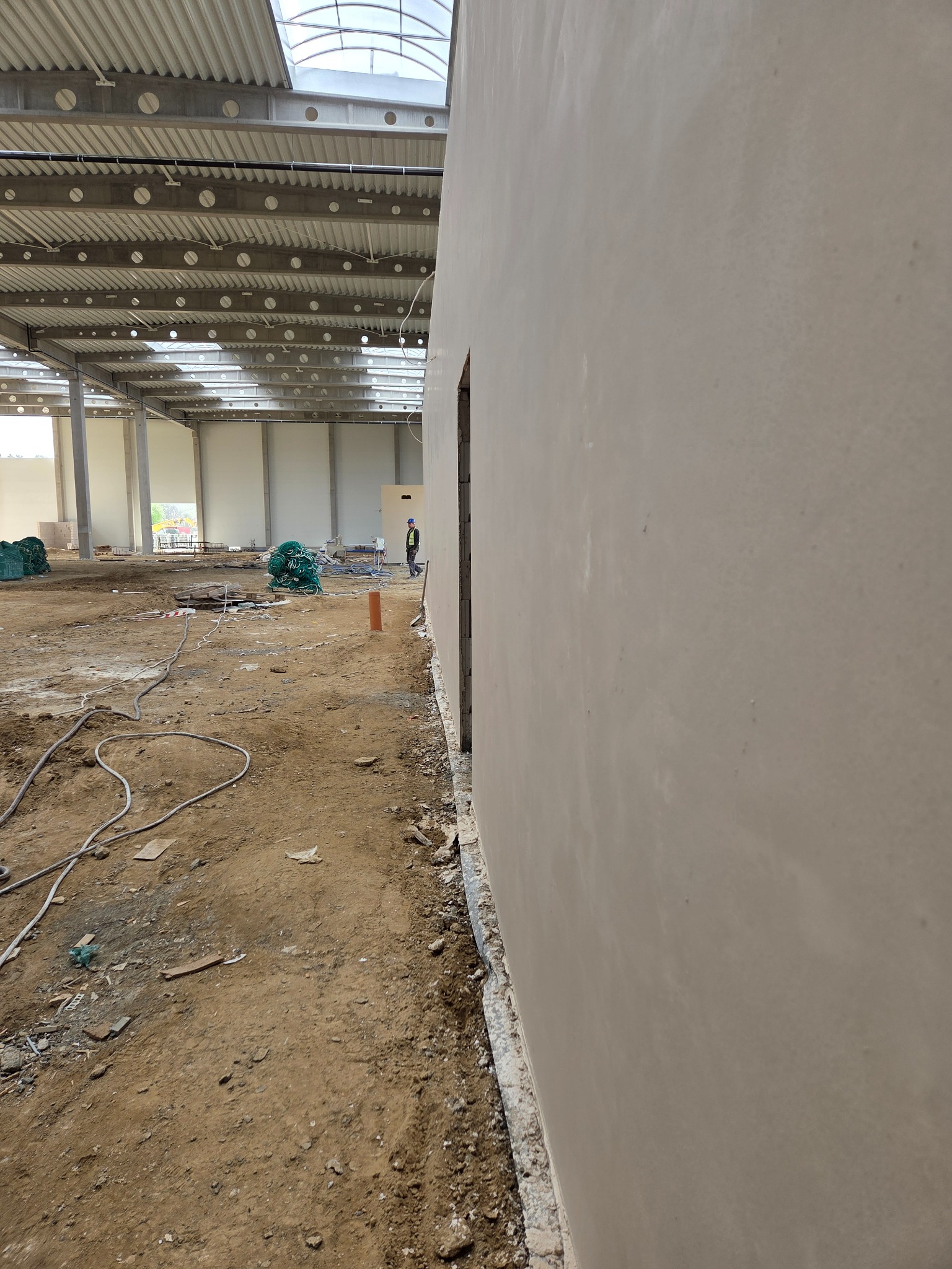 Świeżo otynkowana ściana w hali magazynowej z widocznym fragmentem placu budowy i pracownikiem w tle. Widok z bliska, perspektywa z dołu.