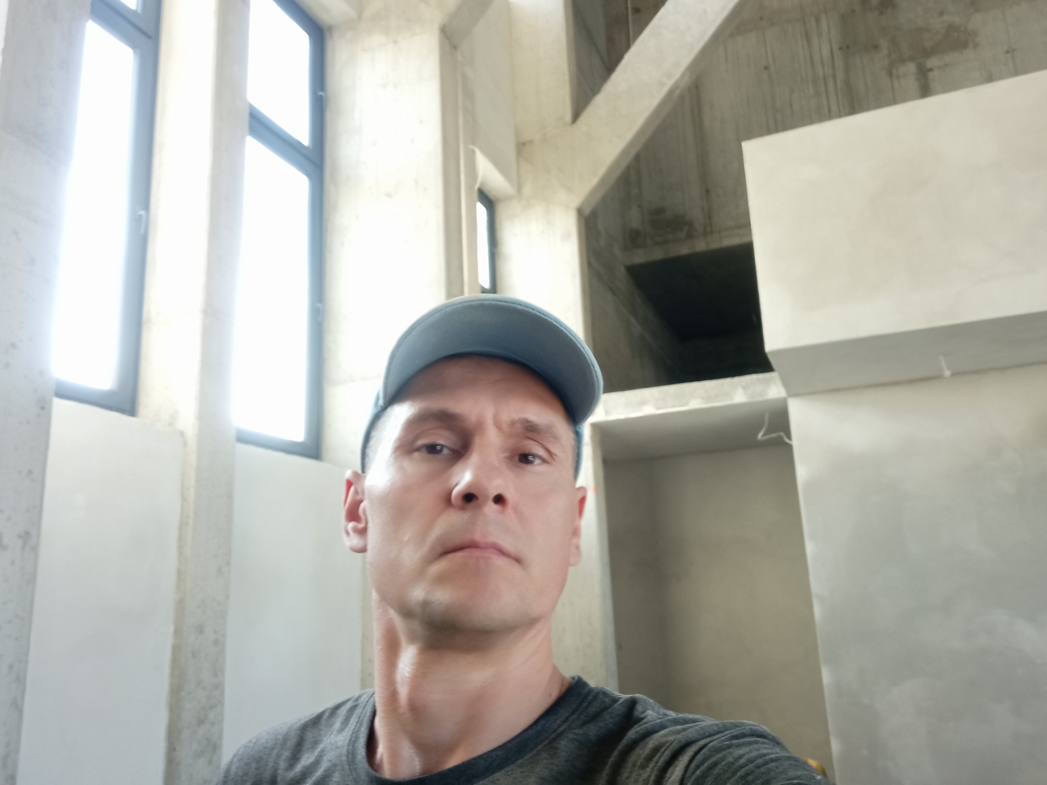 Mężczyzna w czapce na tle surowych ścian po tynkowaniu maszynowym w jasnym wnętrzu. Widoczne okna i betonowa konstrukcja budynku.