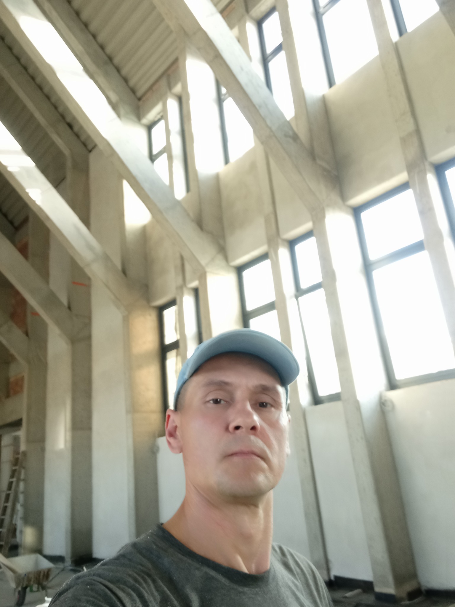 Wnętrze budynku w trakcie prac wykończeniowych; widoczne surowe ściany, okna i elementy konstrukcyjne. Mężczyzna w czapce w kadrze, prawdopodobnie pracownik budowlany.