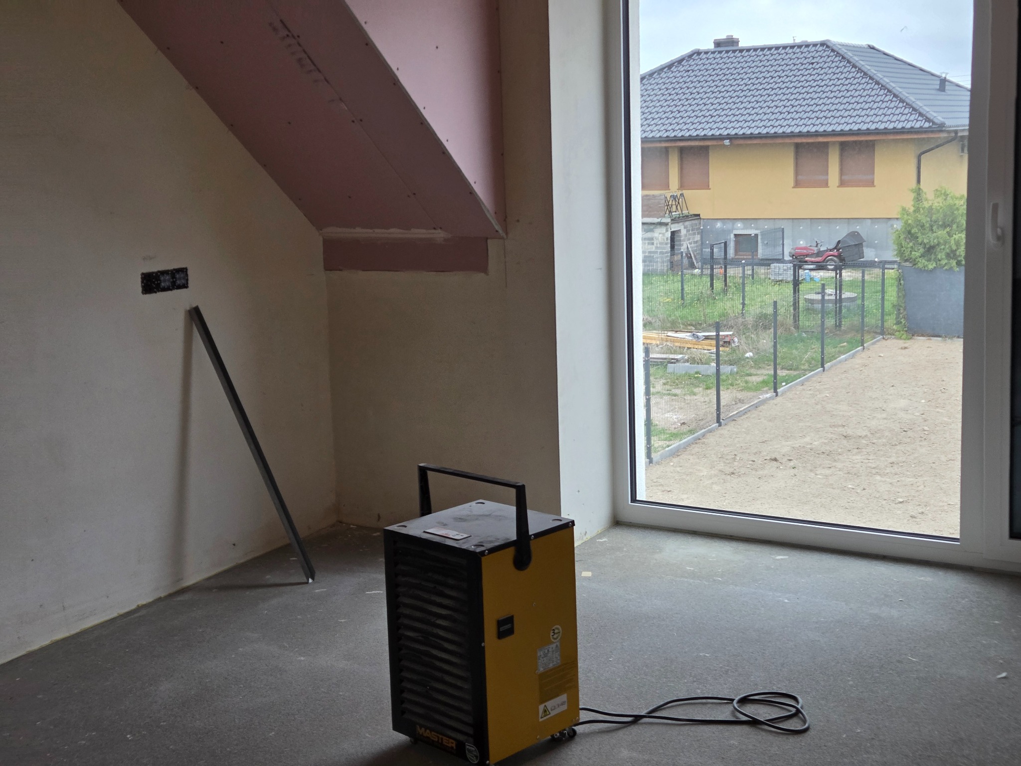 Wnętrze w trakcie prac wykończeniowych: surowe ściany, widoczne przewody elektryczne i nagrzewnica MASTER. W tle okno z widokiem na dom i ogród.