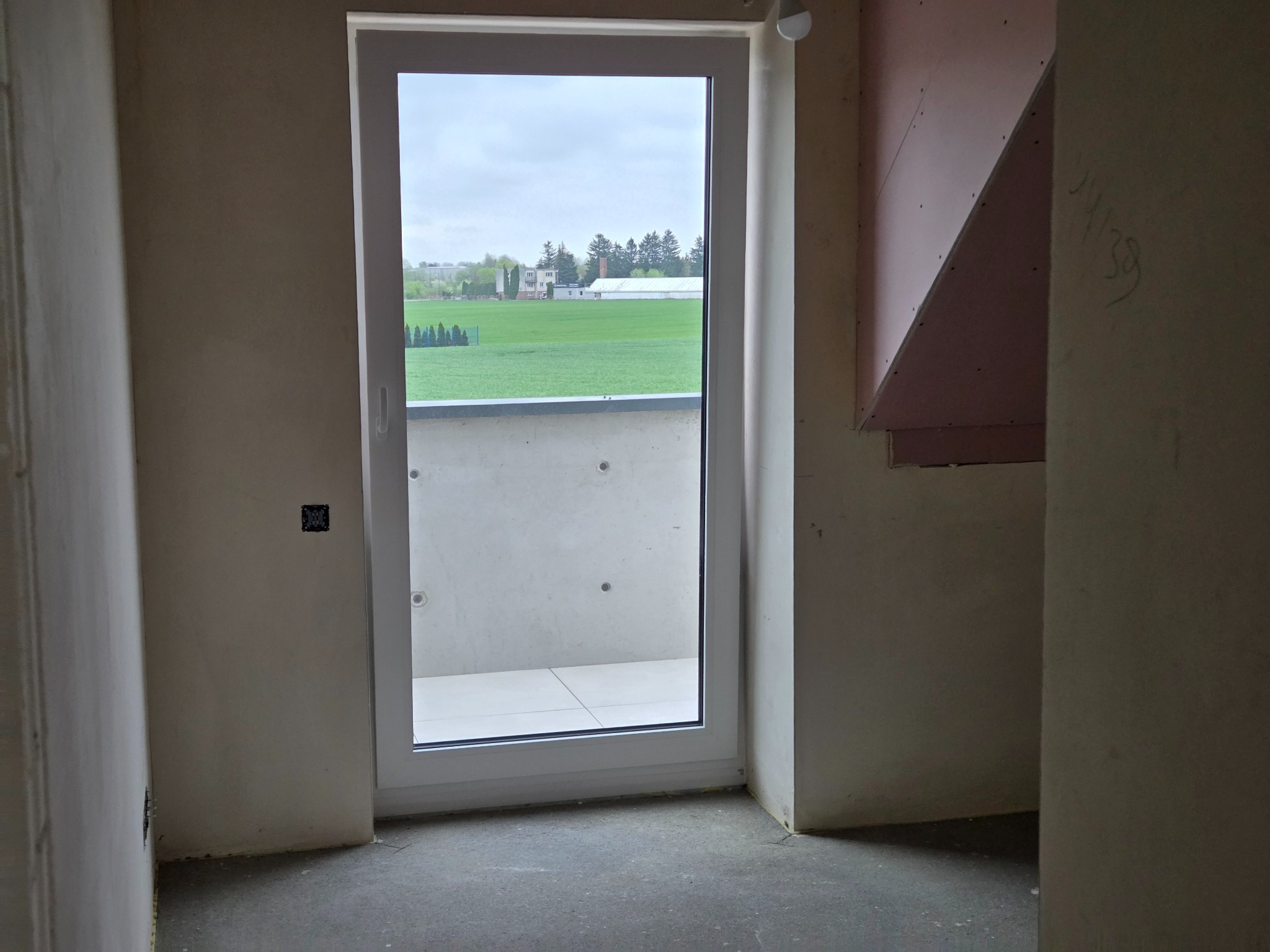 Surowe wnętrze z oknem balkonowym i widokiem na zieleń. Ściany przygotowane do tynkowania, widoczne instalacje elektryczne i fragmenty płyt kartonowo-gipsowych.