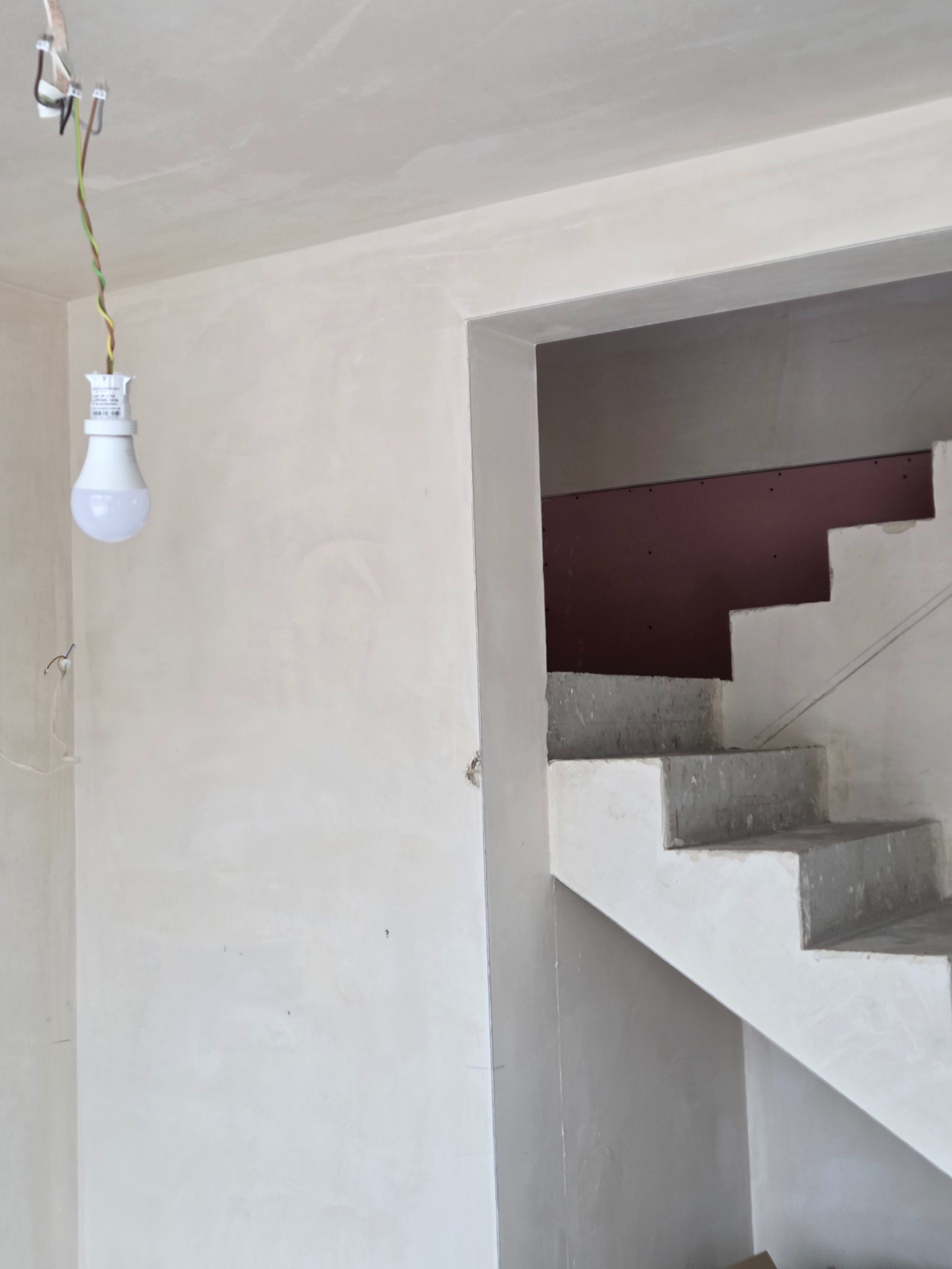 Ściana po nałożeniu gładzi gipsowej, widoczne schody w stanie surowym i żarówka na kablu. Prace wykończeniowe w nowym budynku.
