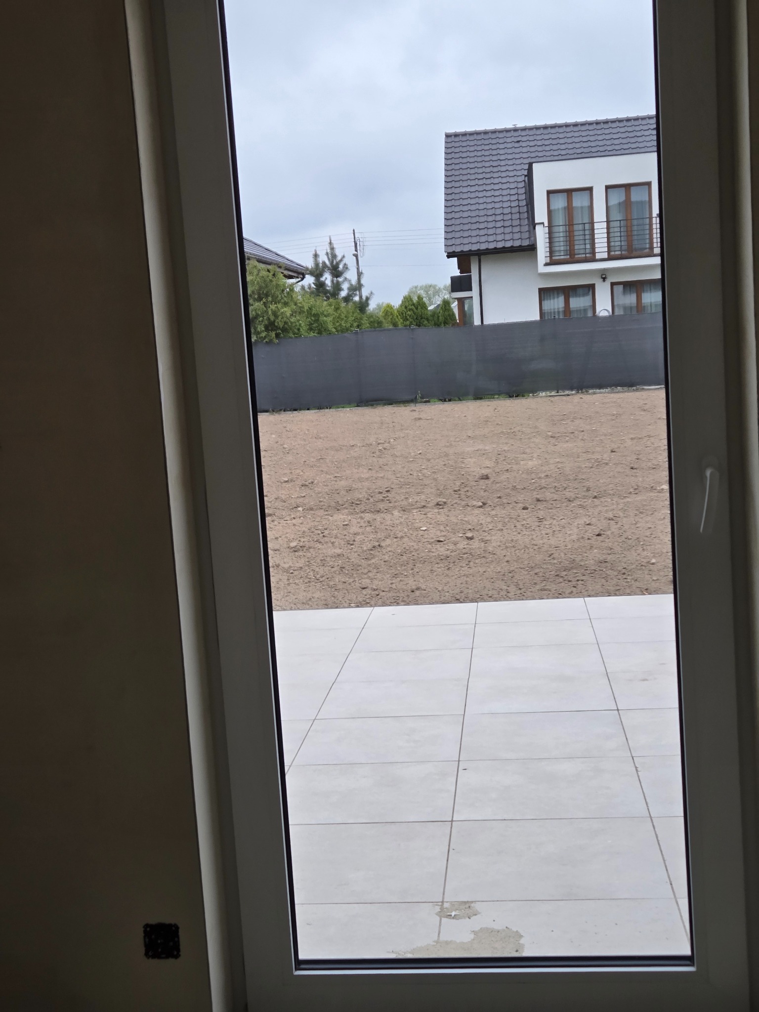 Widok przez okno na przygotowany teren pod trawnik i nowoczesny dom z grafitową dachówką. Jasne płytki na podłodze wewnątrz.