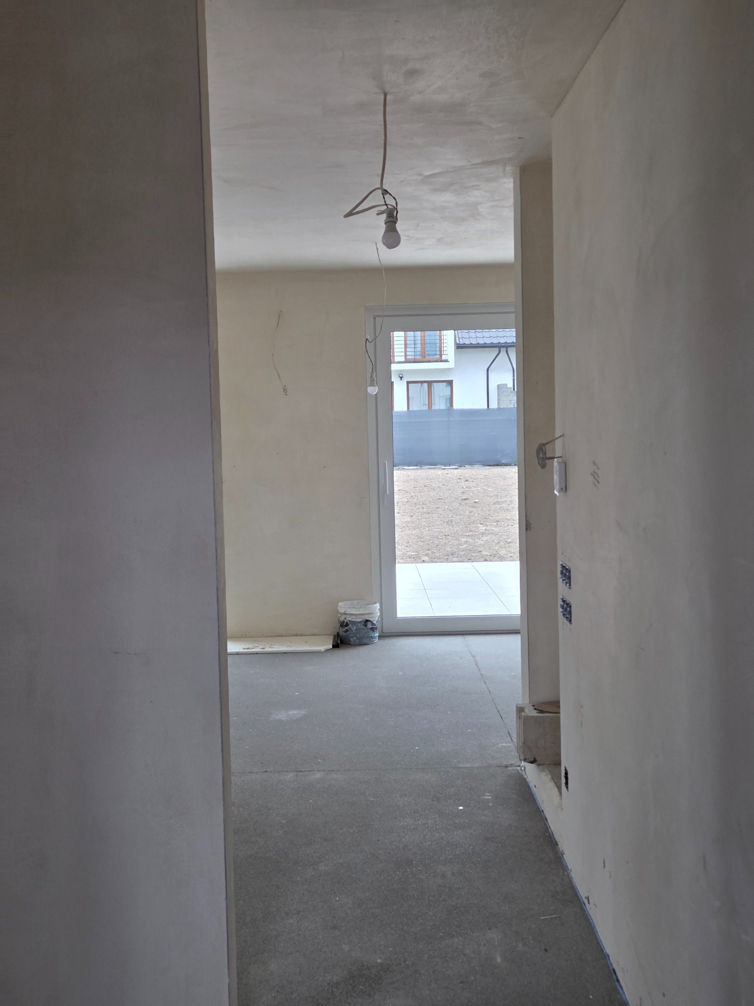 Wnętrze po tynkowaniu maszynowym: gładkie, jasne ściany w surowym stanie, widoczne puszki elektryczne, sufit z wiszącą żarówką, widok na zewnątrz przez przeszklone drzwi.