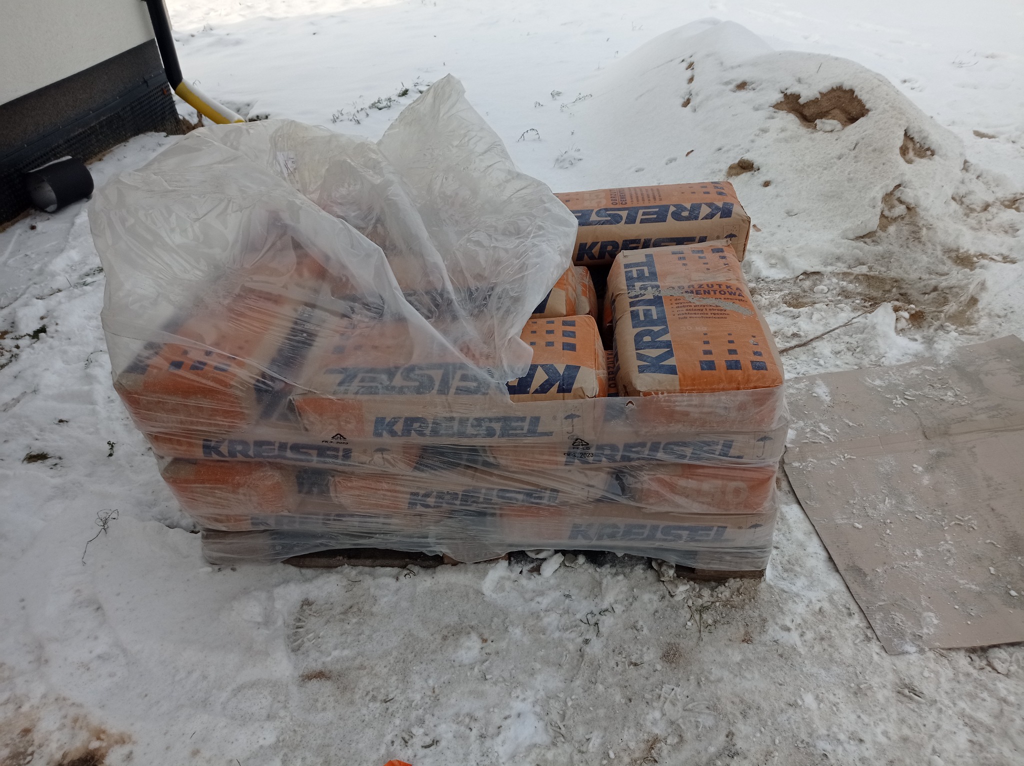 Paleta worków z zaprawą cementową marki Kreisel, owinięta folią, leży na śniegu obok budynku. W tle widać pryzmę piasku.