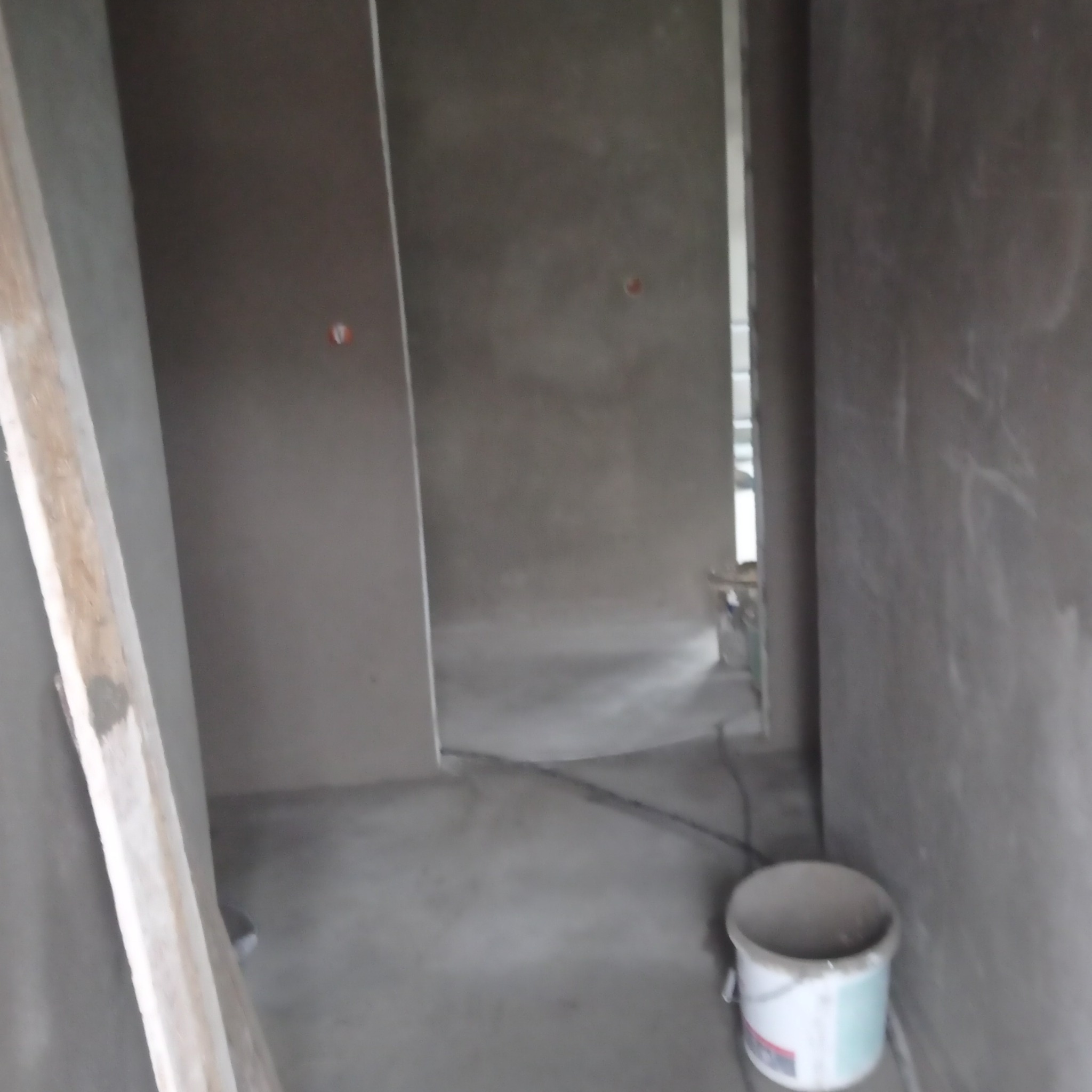 Wnętrze w trakcie prac wykończeniowych: ściany pokryte świeżym, szarym tynkiem, widoczny drewniany profil i wiadro z pozostałościami zaprawy.