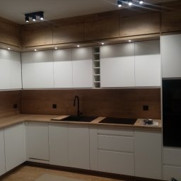 Adam Baczmański - Nowoczesna kuchnia z białymi szafkami i drewnianymi akcentami, czarnym zlewem i płytą indukcyjną, oświetlona punktowymi lampami sufitowymi.