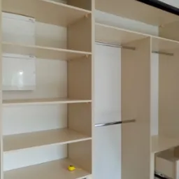 Wnętrze beżowej szafy wnękowej z półkami, drążkami na ubrania i wysuwanymi szufladami, na jednej z półek leży zwinięta żółta miarka.