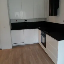 Biała, nowoczesna zabudowa kuchenna z czarnym blatem i minimalistycznym okapem w kształcie walca.