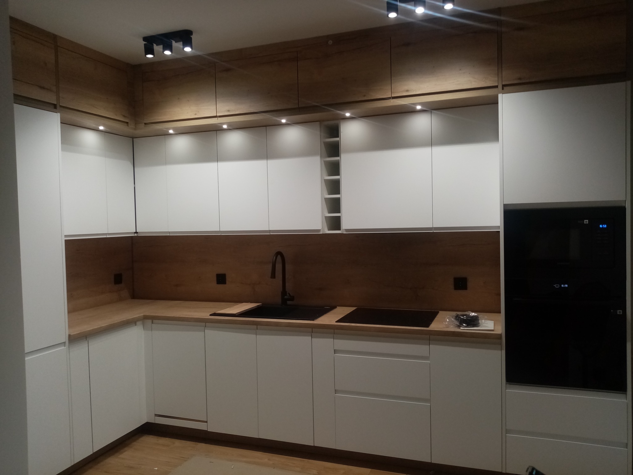 Nowoczesna kuchnia z białymi szafkami i drewnianymi akcentami, czarnym zlewem i płytą indukcyjną, oświetlona punktowymi lampami sufitowymi.