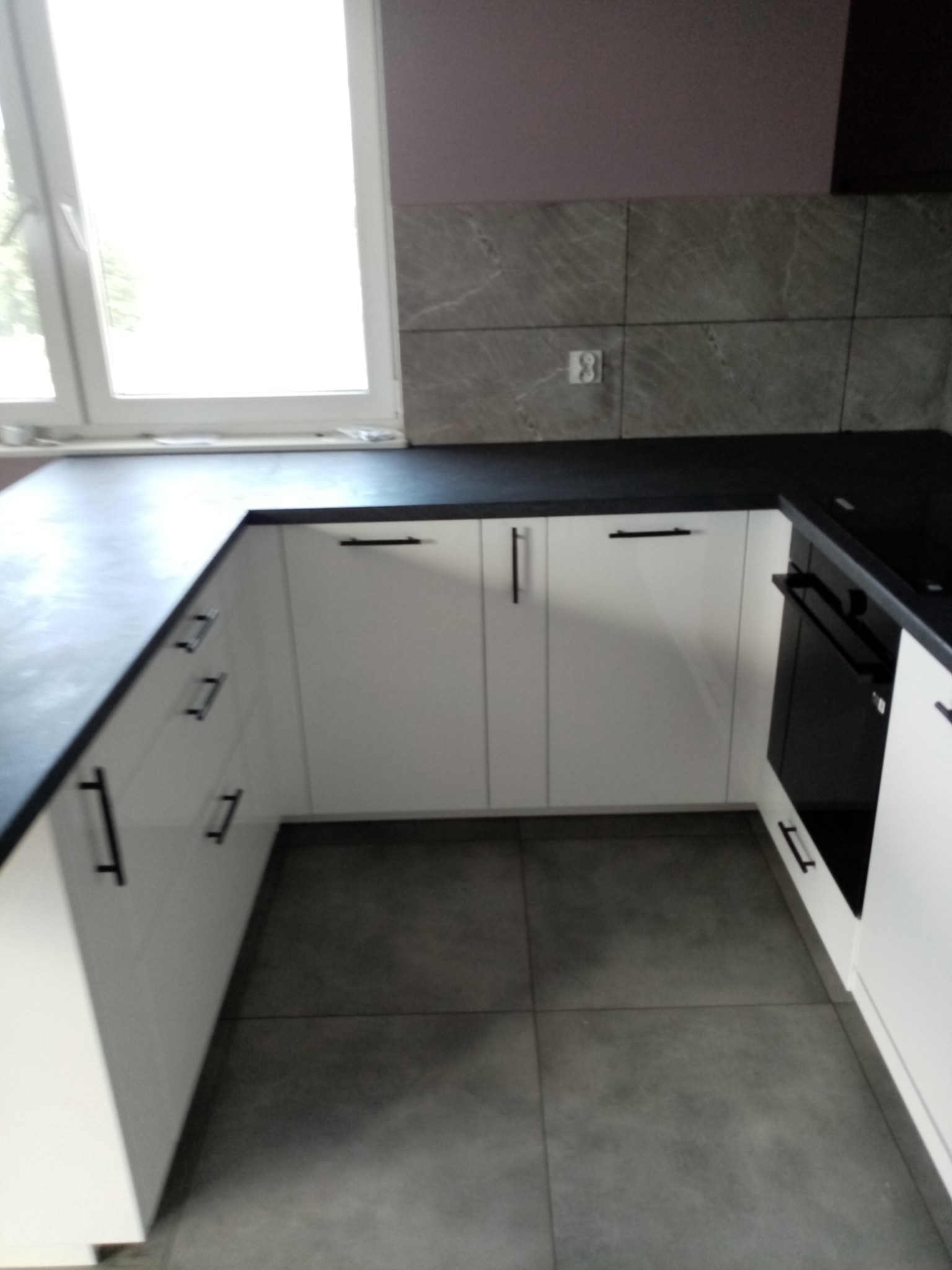 Jasna kuchnia z białymi frontami szafek i czarnym blatem, widok na okno i szare płytki na ścianie.