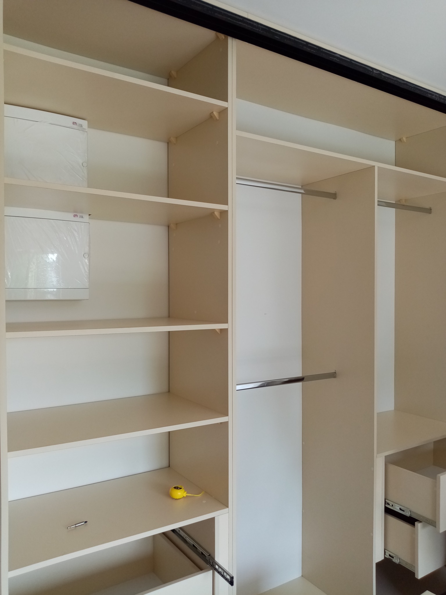 Wnętrze beżowej szafy wnękowej z półkami, drążkami na ubrania i wysuwanymi szufladami, na jednej z półek leży zwinięta żółta miarka.