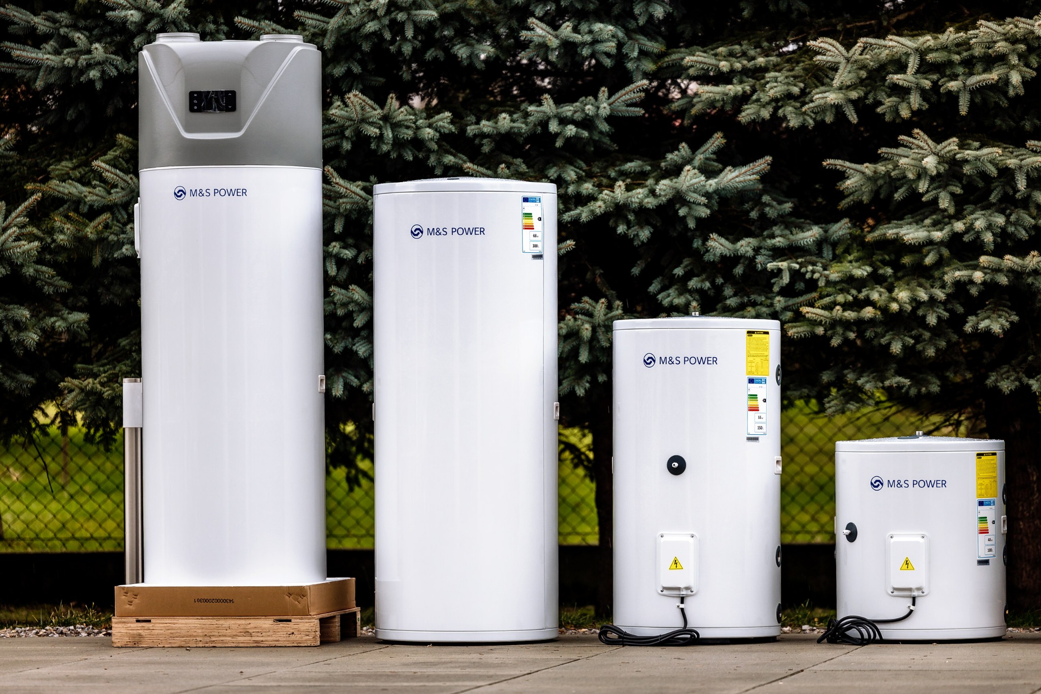 Cztery białe zbiorniki z logo M&S Power, o różnych rozmiarach, stojące obok siebie na betonowej powierzchni przed zielonymi krzewami; największy zbiornik stoi na drewnianej palecie.