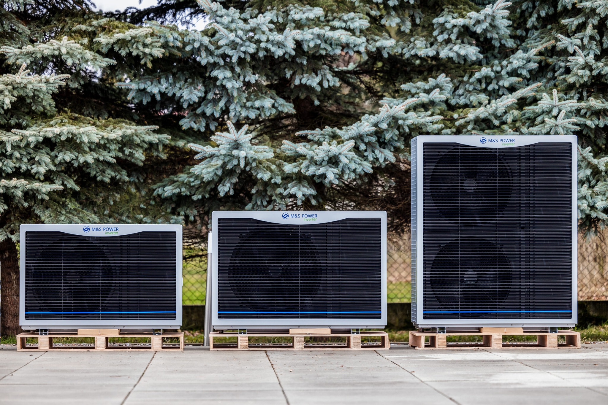 Trzy jednostki różnej wielkości z logo M&S Power na paletach, ustawione na zewnątrz na tle iglastego drzewa.