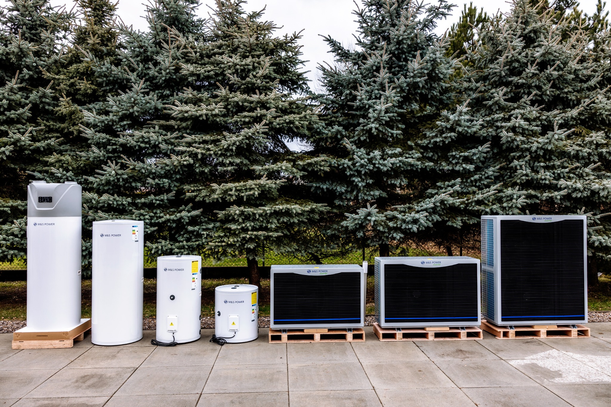 Rząd różnych modeli pomp ciepła marki M&S Power ustawionych na paletach przed rzędem gęstych, zielonych drzew iglastych.