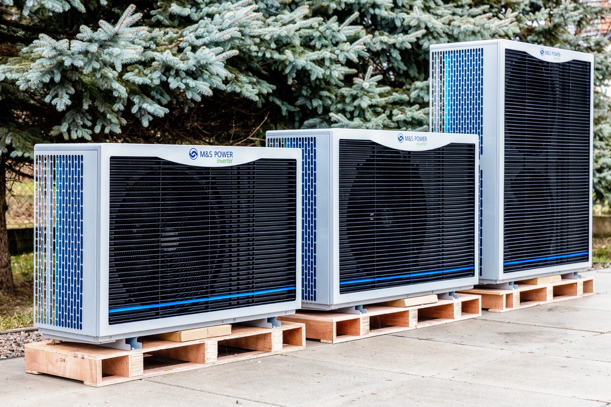 Trzy jednostki zewnętrzne z logo M&S Power ustawione na drewnianych paletach na betonowym podłożu, z iglastym drzewem w tle.
