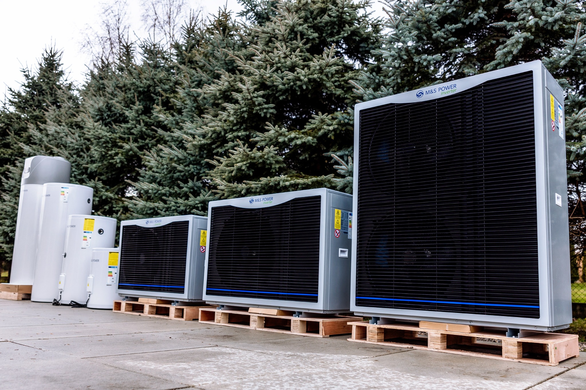 Rząd różnej wielkości jednostek zewnętrznych i zbiorników ciepłej wody użytkowej marki M&S Power, ustawionych na paletach przed rzędem zielonych drzew iglastych.