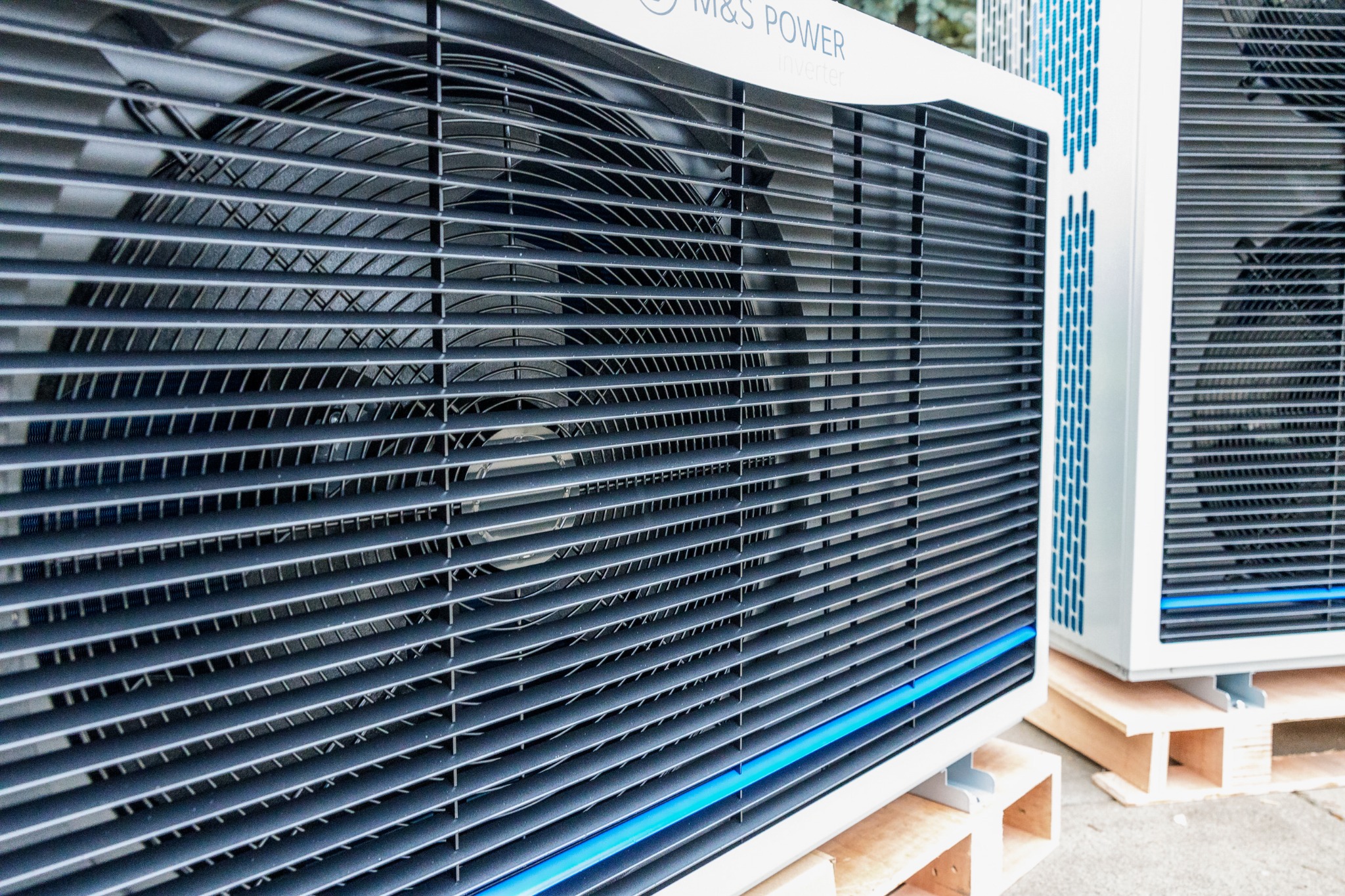 Dwie jednostki zewnętrzne klimatyzacji lub wentylacji, ustawione na drewnianych paletach, z widocznymi żaluzjami i wentylatorami, oświetlone naturalnym światłem.