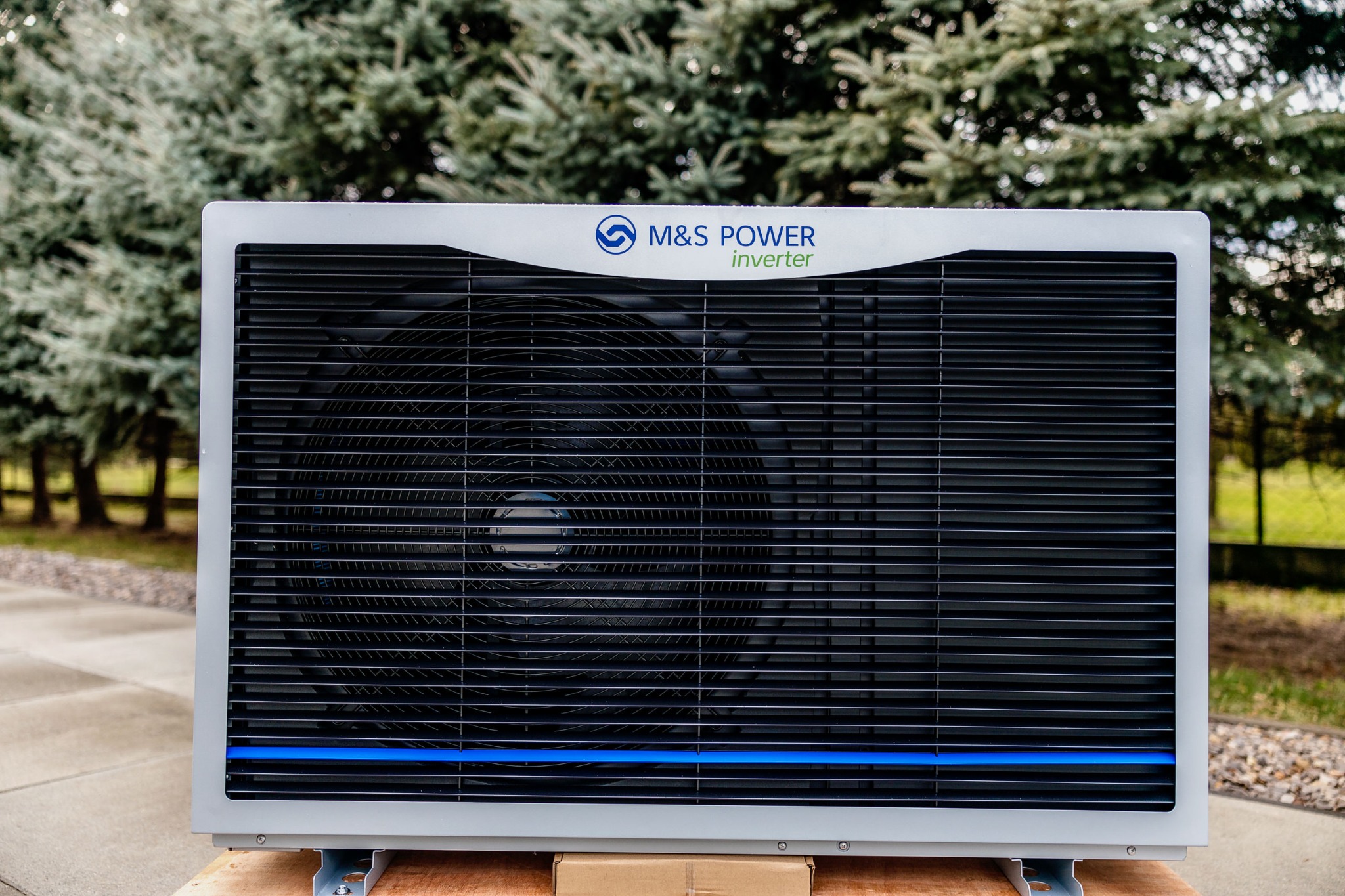 Nowa, nierozpakowana jednostka zewnętrzna z logo M&S Power Inverter, umieszczona na kartonowym pudle na tle iglaków i chodnika.