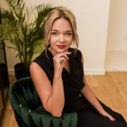 Portret kobiety w czarnym kombinezonie, siedzącej na zielonym, welurowym krześle z ręką podpartą pod brodą, z widocznym złotym pierścionkiem i czerwonymi ustami.