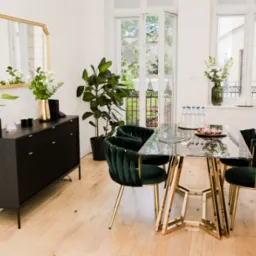 Jasne wnętrze jadalni z marmurowym stołem na złotych nogach, obitymi zielonym aksamitem krzesłami, czarną komodą z lustrem w złotej ramie, kwiatami w wazonie i widokiem na balkon.