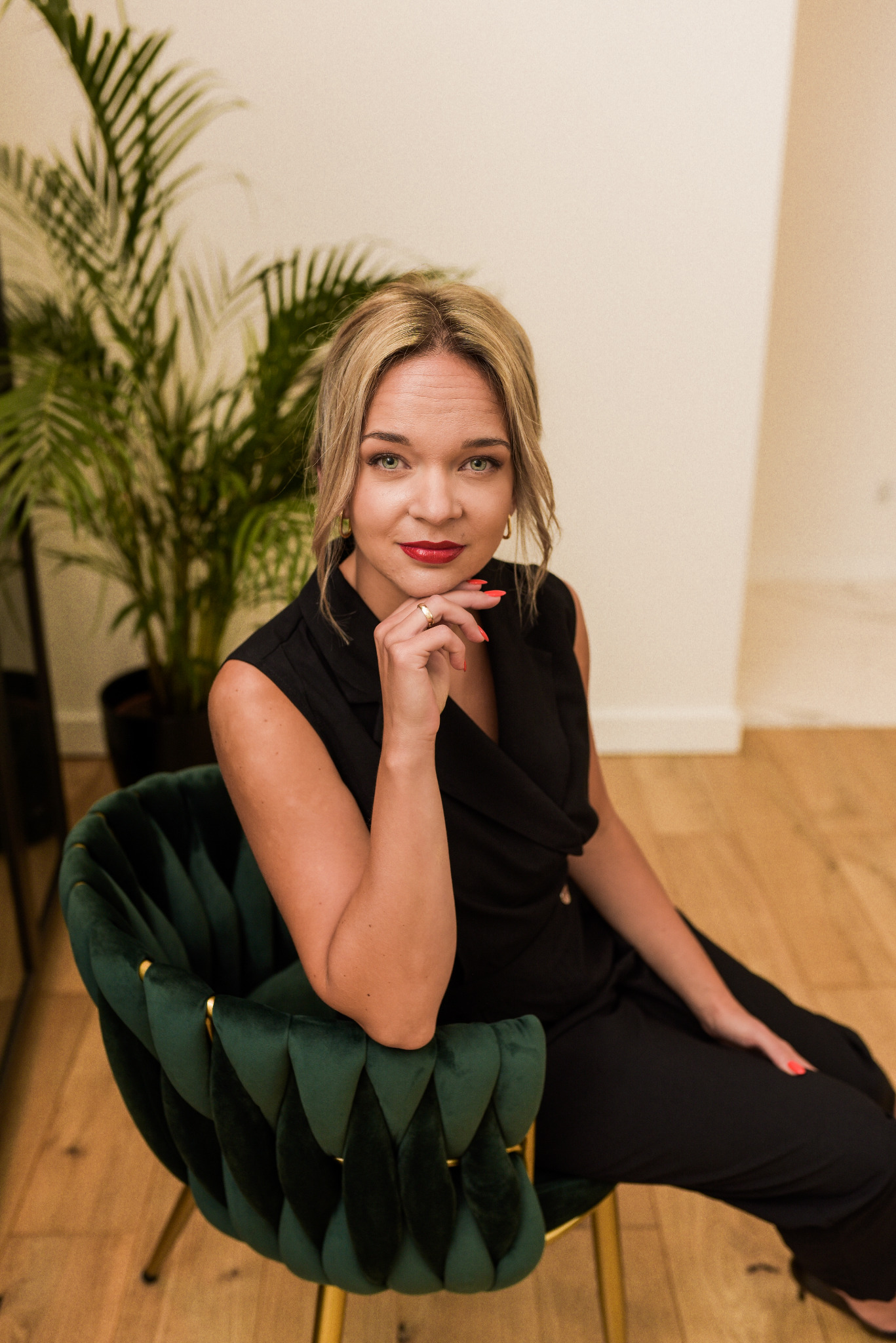 Portret kobiety w czarnym kombinezonie, siedzącej na zielonym, welurowym krześle z ręką podpartą pod brodą, z widocznym złotym pierścionkiem i czerwonymi ustami.
