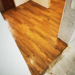 Układanie paneli podłogowych w odcieniach brązu, widoczny surowy betonowy filar i fragment ściany w trakcie wykończenia, częściowo ułożona podłoga w przejściu między pomieszczeniami.