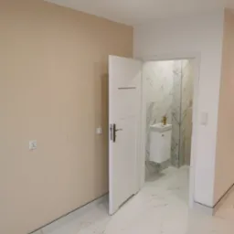 Widok na wykończoną łazienkę z marmurowymi płytkami i złotą armaturą umywalki, widzianą przez otwarte białe drzwi z klamką ze stali nierdzewnej, na podłodze białe płytki imitujące marmur.