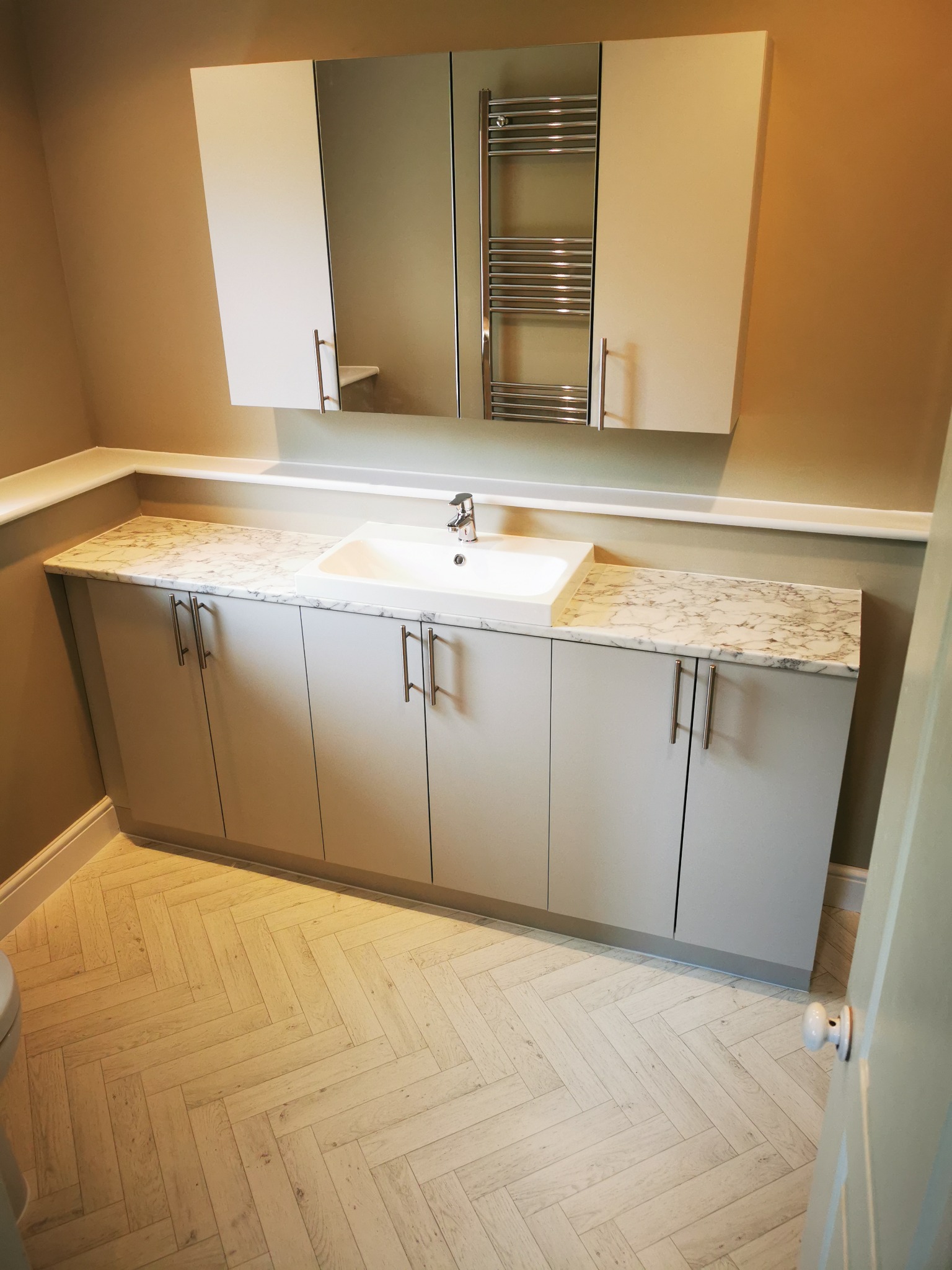 Nowoczesna łazienka z szafką pod umywalką z marmurowym blatem, wiszącą szafką z lustrem i grzejnikiem drabinkowym, podłoga ułożona w jodełkę.