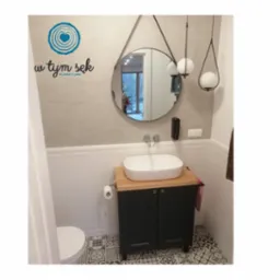 Mała łazienka z umywalką nablatową na szafce z drewnianym blatem, okrągłe lustro na pasku i wiszące lampy kuliste, wzorzysta podłoga, widoczne logo firmy na ścianie.