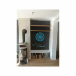 Wnętrze z zabudową ściany: czarna ściana z drewnianymi półkami, biała półka dolna z ułożonym drewnem, obok piec wolnostojący z drewnianą obudową i logo firmy na ścianie.