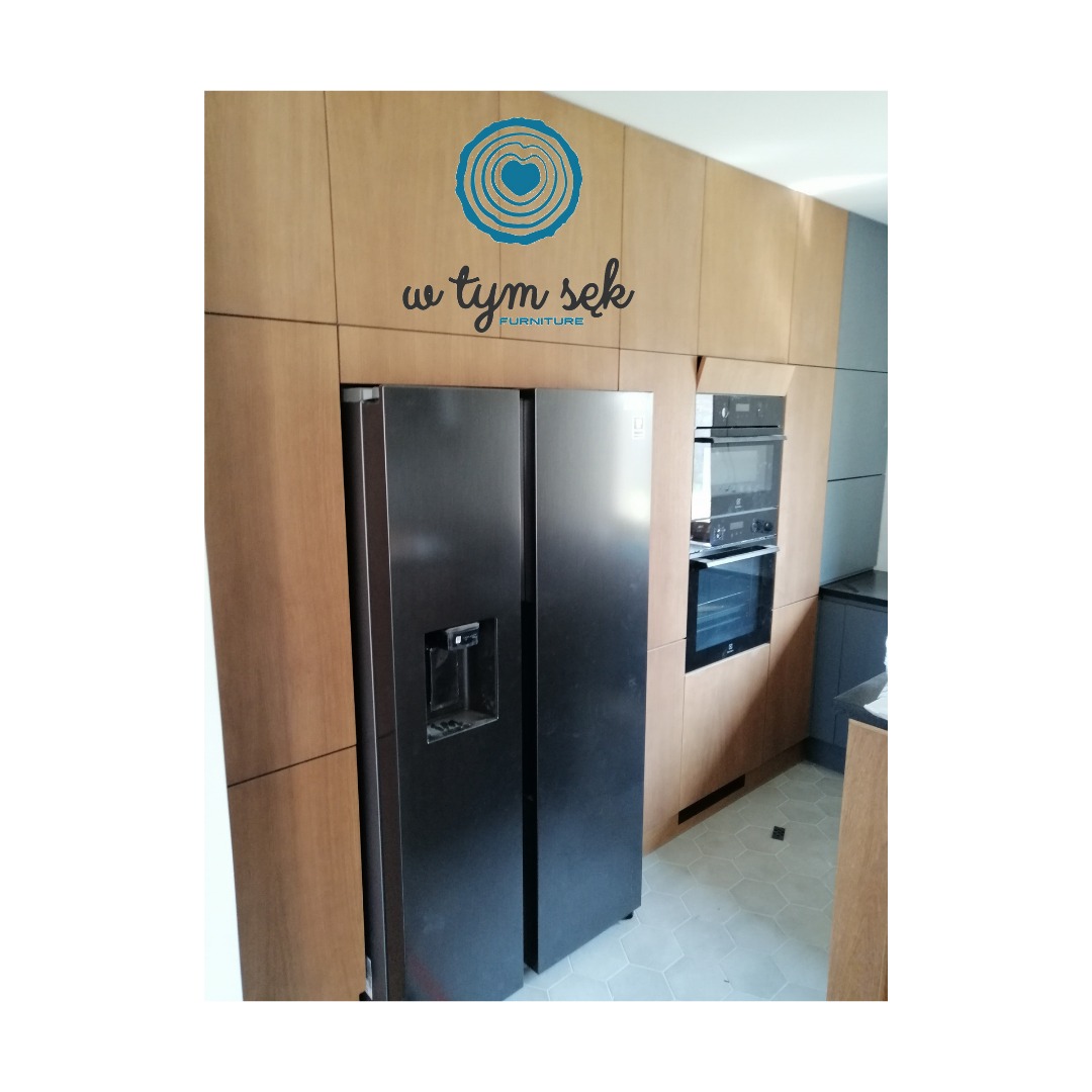 Nowoczesna kuchnia z zabudową drewnianą, obejmującą lodówkę side-by-side i piekarnik, z logo firmy meblarskiej na górnej szafce.