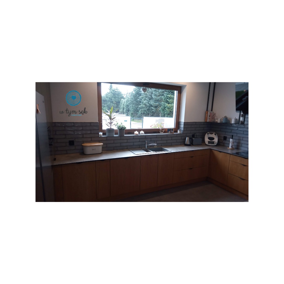 Nowoczesna kuchnia z drewnianymi frontami szafek, blatem w odcieniach szarości i oknem z widokiem na drzewa; nad blatem dekoracyjny napis na ścianie.