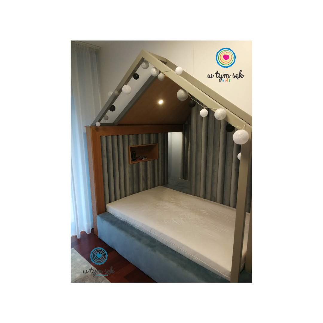 Łóżko dziecięce w kształcie domku z szarą tapicerowaną ścianą, drewnianą ramą i oświetleniem z kul, widoczne logo firmy w rogu.