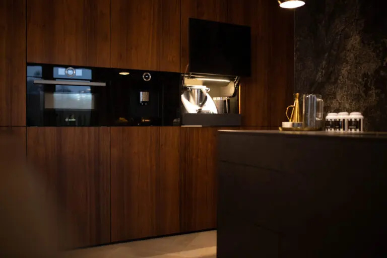 Nowoczesna kuchnia z drewnianymi frontami, zabudowany piekarnik i ekspres do kawy, wysuwana szafka z robotem kuchennym, ciemny blat i akcesoria kuchenne.