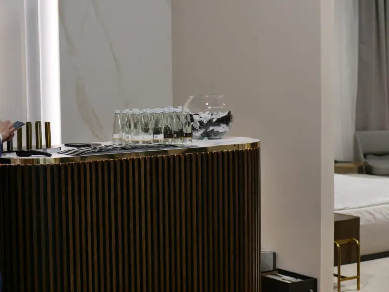 Nowoczesna zabudowa barowa z pionowymi lamelami w kolorze ciemnego drewna, zwieńczona białym blatem z butelkami wody i szklanym naczyniem z dekoracyjnymi elementami, widoczna częściowo sypialnia...