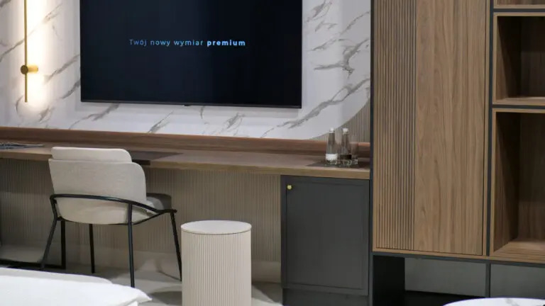 Nowoczesny pokój hotelowy z biurkiem z blatem drewnianym, krzesłem z metalową ramą, szafką z ciemnoszarymi drzwiczkami i telewizorem na ścianie z marmurowym wzorem.