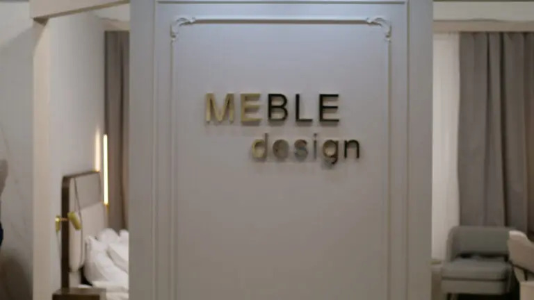 Ściana z napisem 'MEBLE design' w złotych i czarnych literach, widok na aranżację wnętrza z łóżkiem i krzesłem w tle, Siepraw.