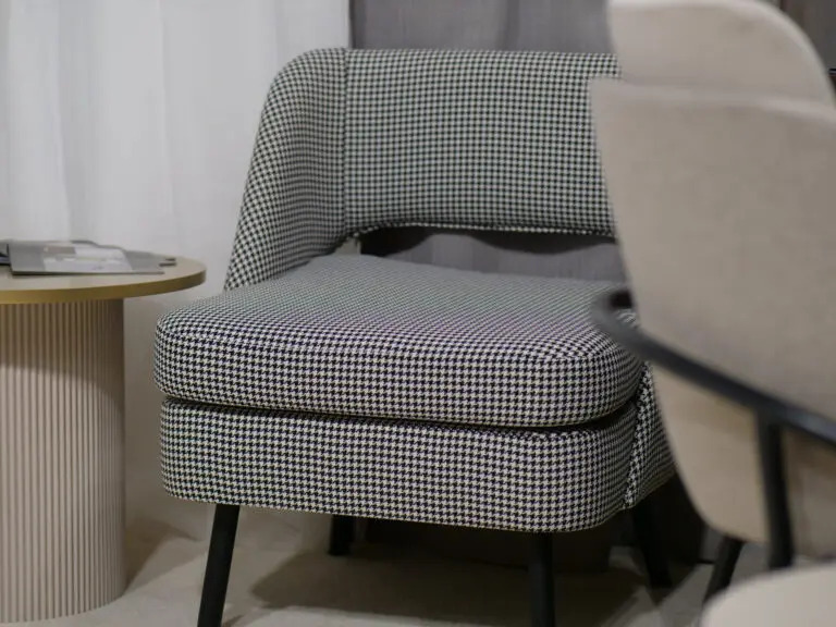 Tapicerowane krzesło w pepitkę z czarnymi metalowymi nogami, obok okrągły stolik z blatem w kolorze złota i żłobioną kolumnową podstawą.