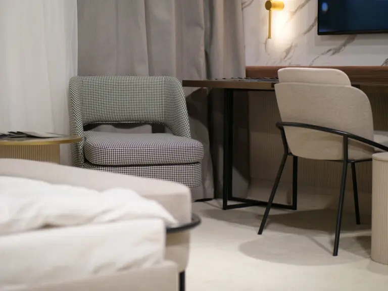 Nowoczesny pokój z dwoma krzesłami: jedno w pepitkę, drugie beżowe z czarną ramą, biurko z czarnymi nogami i marmurowa ściana z telewizorem i złotym kinkietem.