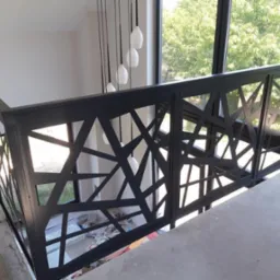 Czarna balustrada z geometrycznym wzorem na tle okna z widokiem na zieleń, w trakcie montażu na betonowej powierzchni