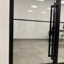 Szklane drzwi biurowe z czarną, metalową ramą i uchwytami, widok na wnętrze biura z biurkami i krzesłami.