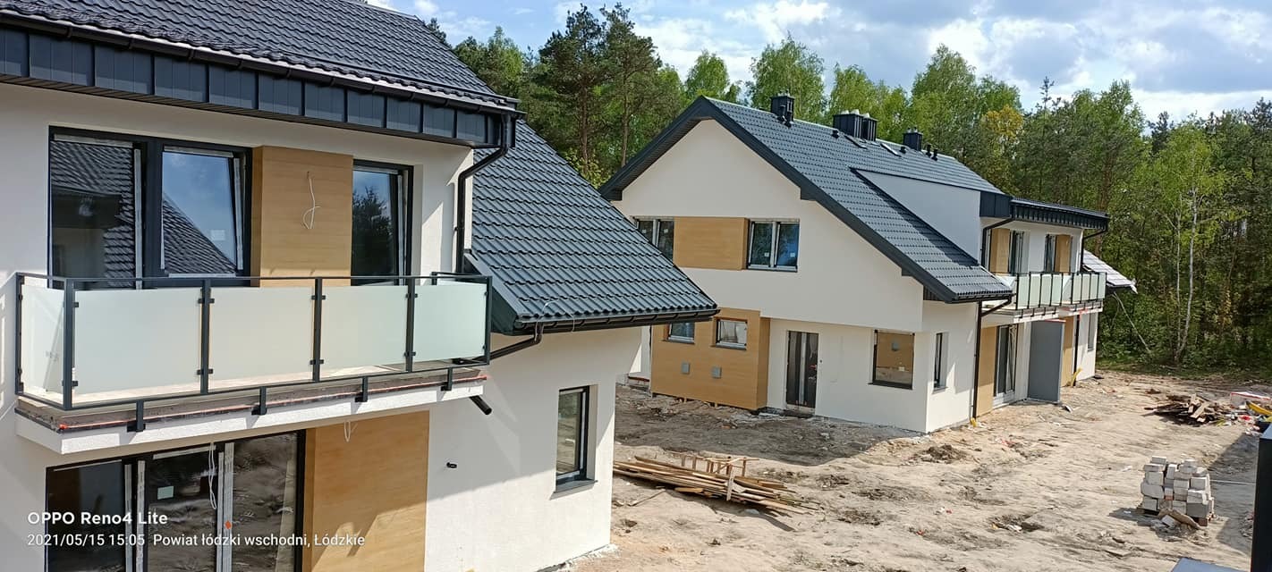 Dwa nowoczesne domy jednorodzinne z balkonami z balustradami ze szkła mrożonego i czarnymi ramami, widoczne drewniane elementy elewacji, teren wokół w trakcie prac wykończeniowych.