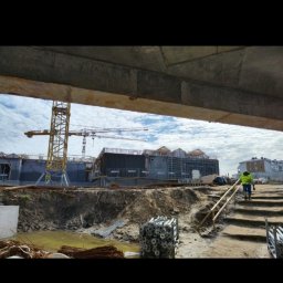 Robert Łaszczewski - Plac budowy z widocznymi elementami konstrukcyjnymi, żurawiami i pracownikiem w odblaskowej kamizelce w tle, z ziemią i materiałami budowlanymi na pierwszym planie, ujęcie spod betonowej konstrukcji.