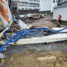 Robert Łaszczewski - Plac budowy z rusztowaniami przy budynku, widoczne niebieskie rury, pracownik w kamizelce odblaskowej z narzędziami i pomarańczowy kabel na ziemi.