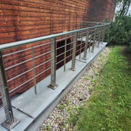 Robert Łaszczewski - Zewnętrzna balustrada ze stali nierdzewnej, zamontowana przy drewnianej elewacji budynku, z widocznym fragmentem trawnika i kamiennego podłoża.