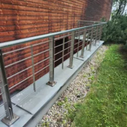 Zewnętrzna balustrada ze stali nierdzewnej, zamontowana przy drewnianej elewacji budynku, z widocznym fragmentem trawnika i kamiennego podłoża.