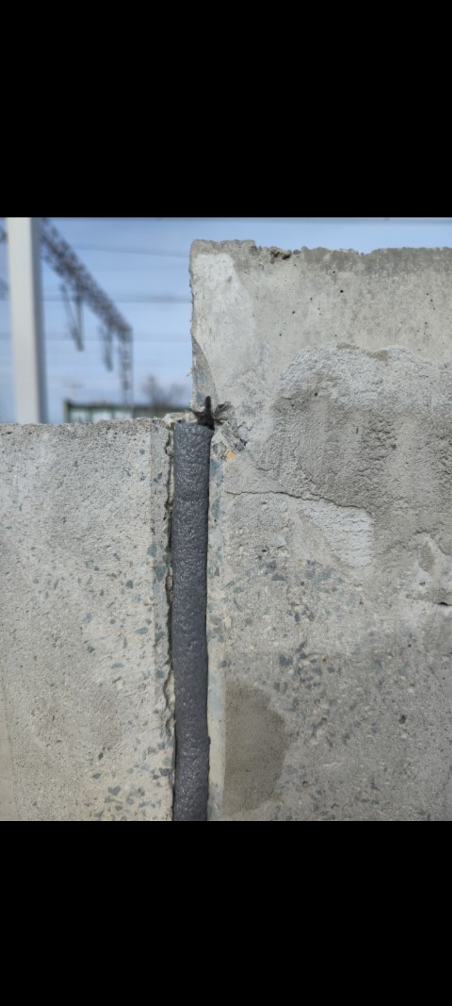 Szczelina dylatacyjna wypełniona szarą masą uszczelniającą pomiędzy betonowymi prefabrykatami, widoczne ślady napraw na powierzchni betonu, w tle słupy wysokiego napięcia.