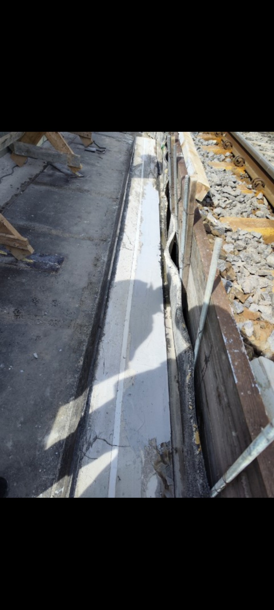 Szczelina między betonowym chodnikiem a drewnianą obudową torowiska, wypełniona szarym materiałem z widocznymi pęknięciami i uszkodzeniami, w tle żwir i szyny kolejowe.